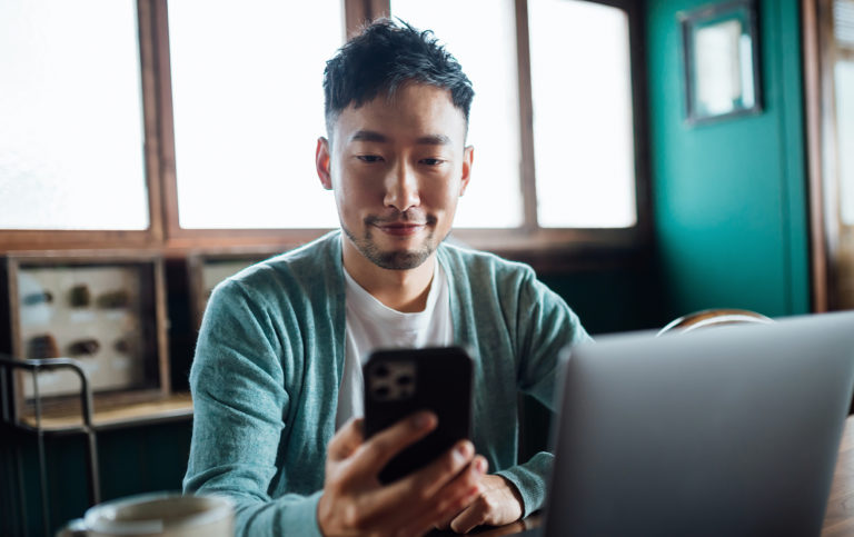 Confident young Asian man looking at smartphone while working on laptop computer image