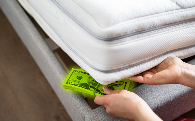 A person storing a stack of $50 bills underneath a mattress.