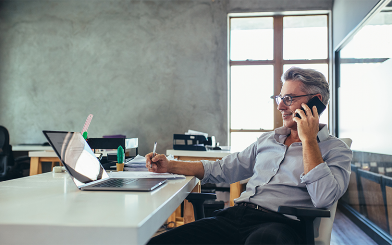 Middle-aged man sits at desk while talking on the phone.